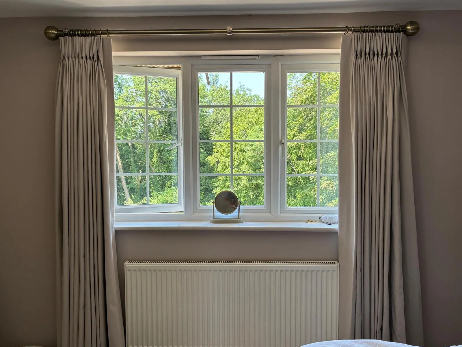 Pale pinch-pleat curtains on an antique-brass pole framing a cottage window and garden view